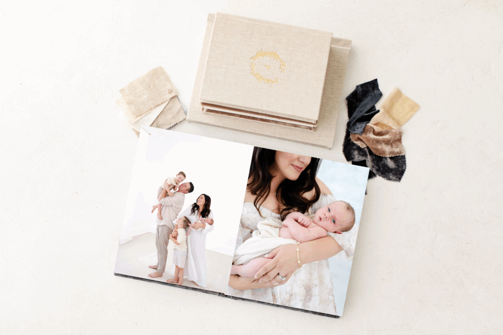 Heirloom album with linen cover and gold foil emblem, opened to portraits of a newborn and family in soft, natural light.